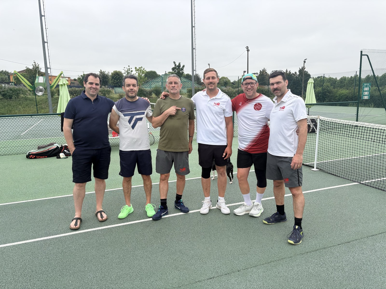 Six joueurs adultes du club posant devant le filet sur un court extérieur, dont trois portent le polo rouge et blanc du club