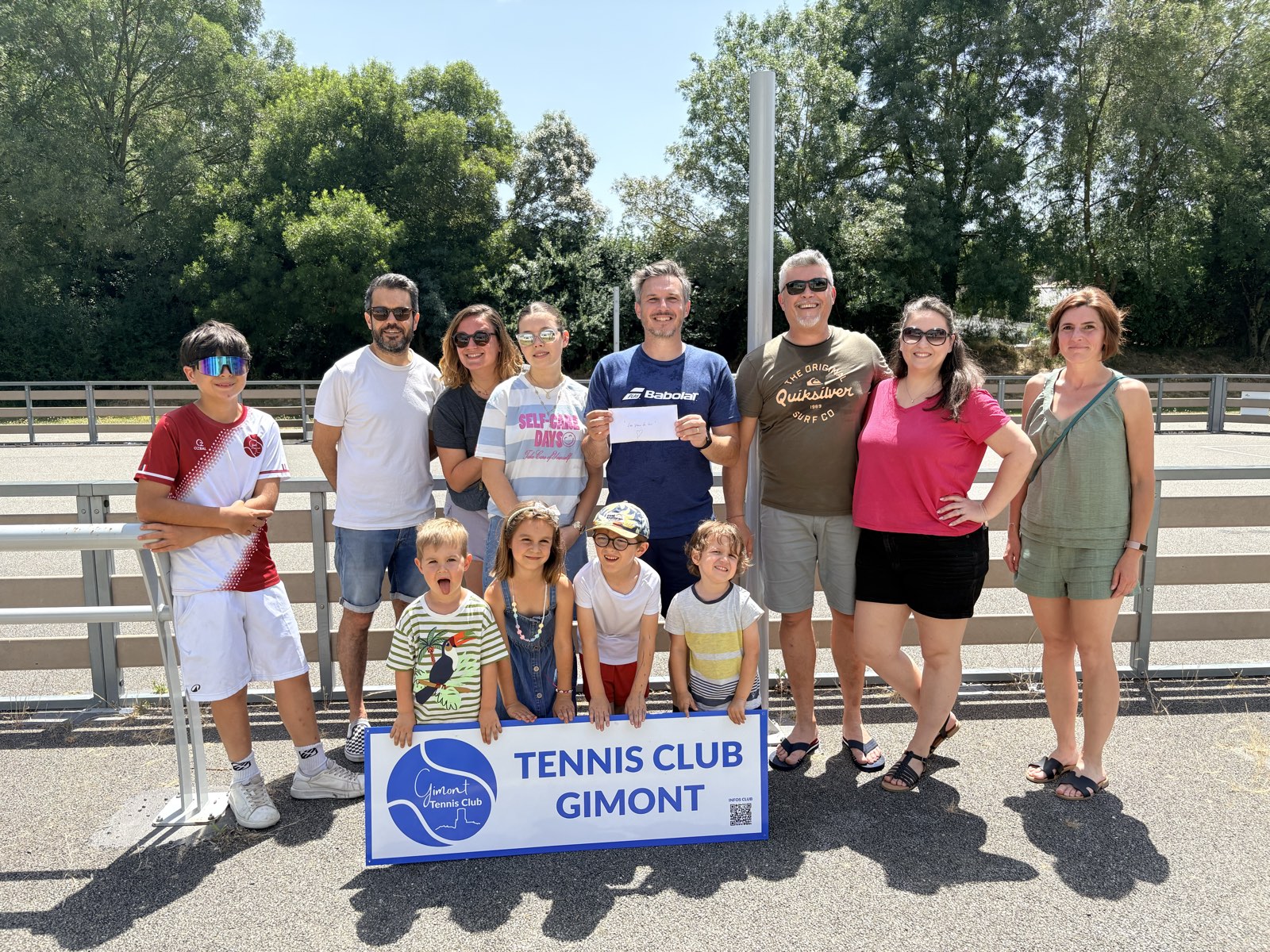 Les joueurs et les familles du club entourent Nathanaël et sa famille lors de la remise des bénéfices du tournoi à l'association Les yeux de Lou, devant la banderole du Tennis Club Gimont